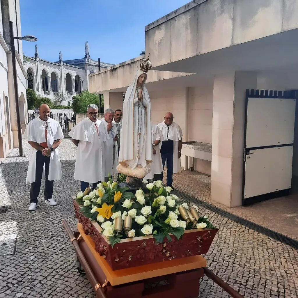 A imagem retrata uma estátua de Nossa Senhora de Fátima posicionada sobre um pedestal decorado com flores. A estátua, vestida com um manto branco, segura um terço. Ao redor dela, quatro indivíduos vestindo roupas religiosas brancas estão presentes. O cenário de fundo mostra uma estrutura arquitetônica com colunas e arcos, possivelmente parte de uma igreja ou outro edifício religioso. O céu é claro e azul, indicando que a foto foi tirada durante o dia em condições climáticas favoráveis.