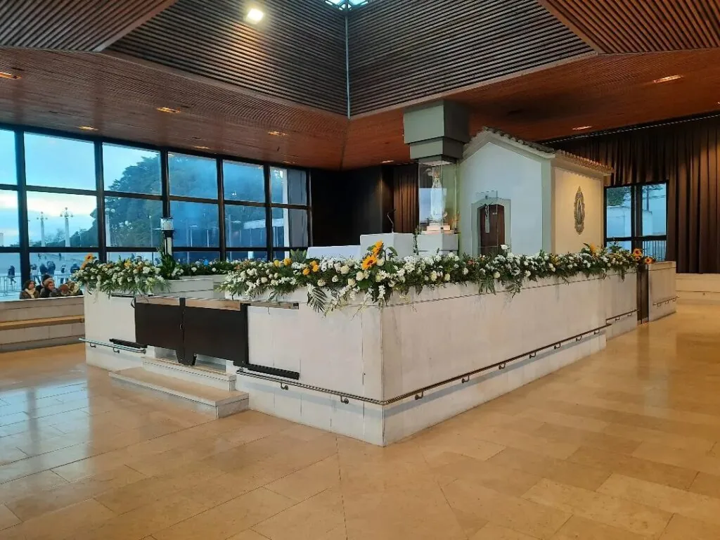 A imagem retrata um altar em uma igreja, adornado com flores brancas e verdes. O altar é feito de um material claro, possivelmente mármore ou uma pedra similar. Atrás do altar, há uma pequena capela religiosa protegida por vidro e fixada na parede. O teto acima é de madeira, com padrões lineares e luminárias embutidas que iluminam o espaço abaixo. Grandes janelas de vidro estão visíveis ao fundo, permitindo a entrada de luz natural; o lado de fora parece ser o amanhecer ou o entardecer devido à iluminação suave. O piso é composto por lajes de pedra polida, refletindo parte das luzes do ambiente.