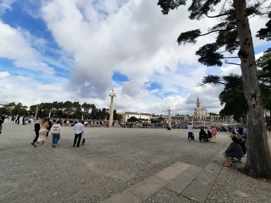Pessoas caminhando pela Esplanada do Santuário de Fátima sob um céu com nuvens brancas. A Basílica da Santíssima Trindade e a Basílica de Nossa Senhora do Rosário de Fátima estão visíveis ao fundo